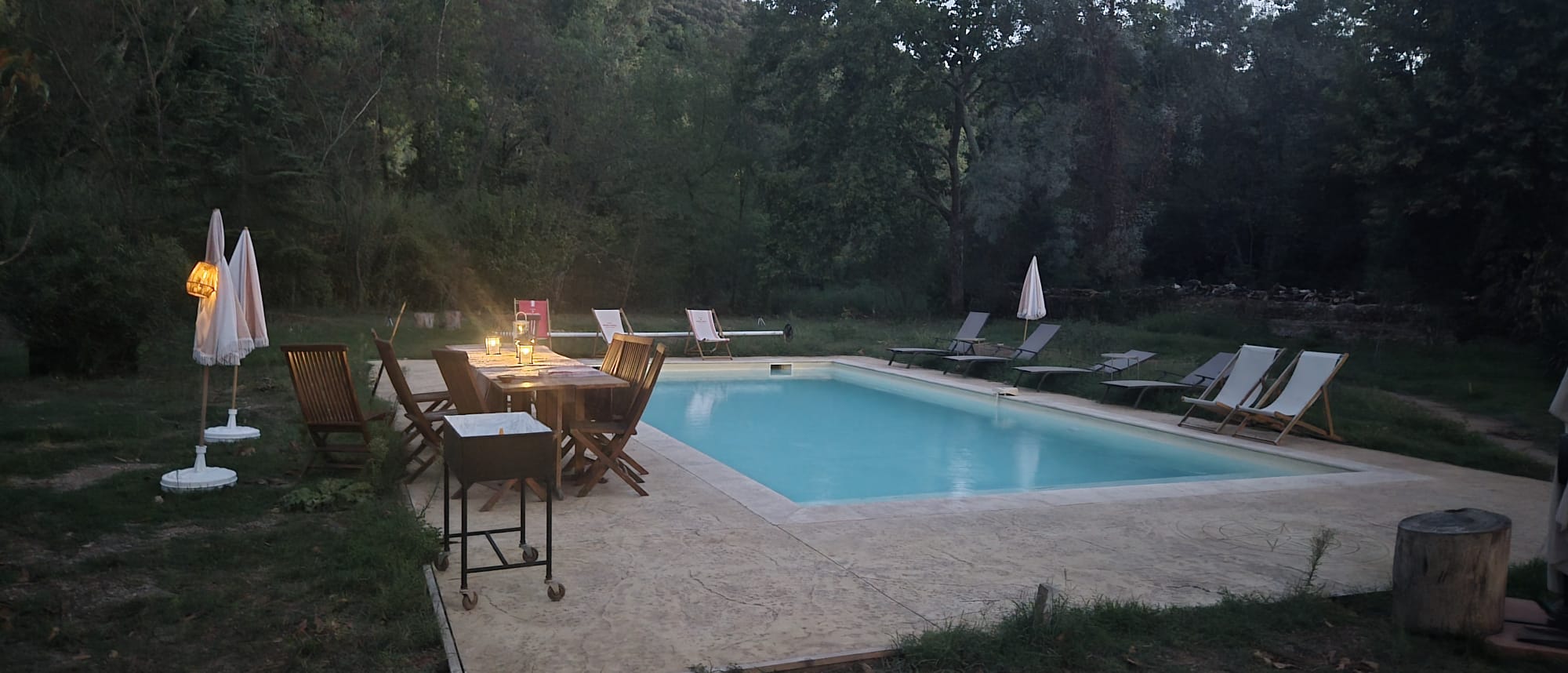 La piscine au crépuscule, avec une table éclairée et des transats au bord du parc