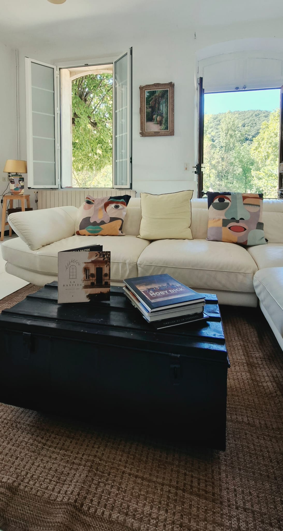 Le salon de LA VILLAMARA, canapé et livres face aux fenêtres ouvertes sur les arbres